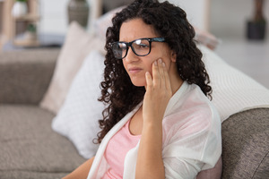 Woman with TMJ disorder sitting on couch rubbing jaw in pain